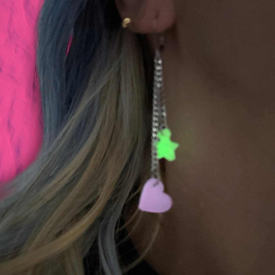 A closeup of a woman's earlobe in low light showcases a dangly glow-in-the-dark pink heart and green star earring.