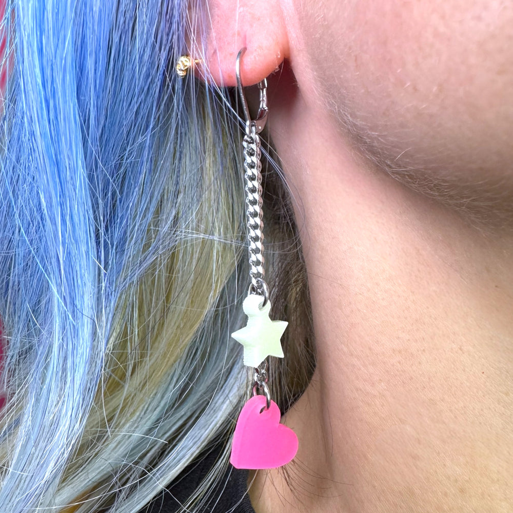 Close up of a woman's earlobe wearing a linked glow-in-the-dark pink heart and light green star dangle earring with stainless steel findings.