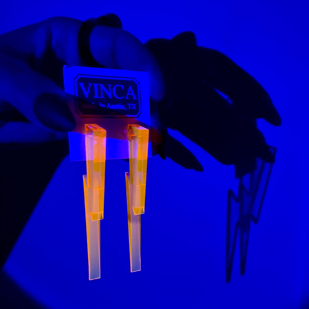 A hand holds a Vinca branded card with transparent pink Lightning Bolt earrings against a blue background.