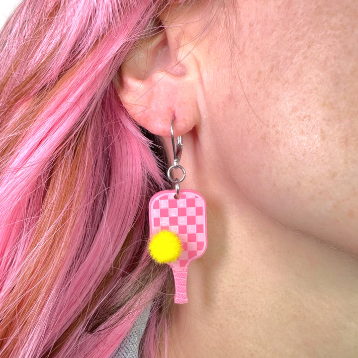 A woman with pink hair wears a pink pickleball paddle dangle earring with yellow pompom detail.