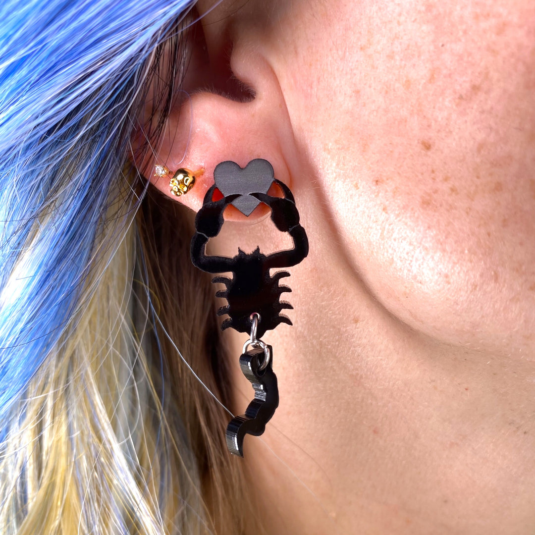 A woman with medium length blue hair wears a black scorpion holding a heart earring. 