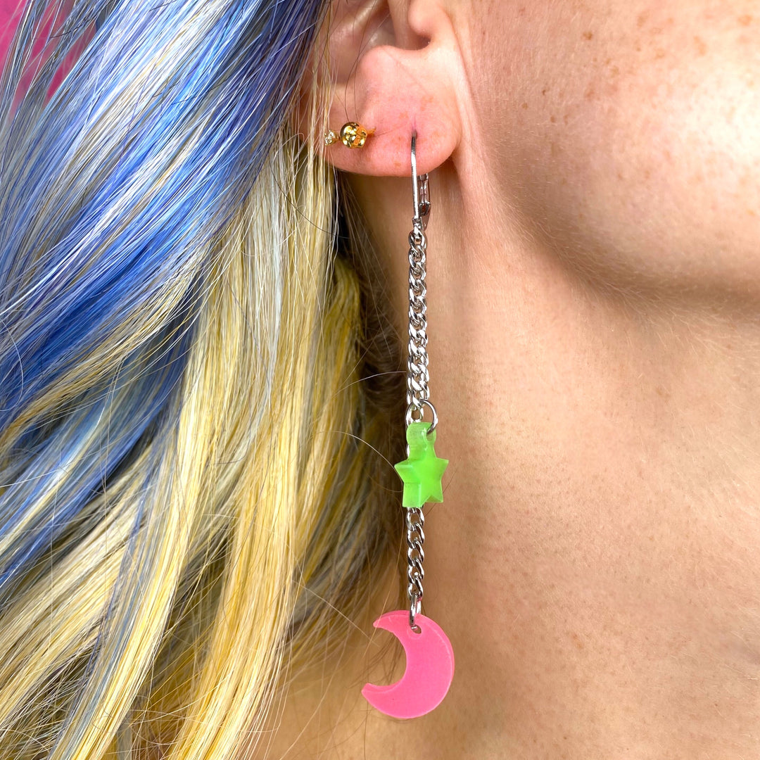 Our limited edition glow-in-the-dark Moon and Star stainless steel dangle earrings in pink & green are worn by a woman with blue highlighted hair. 