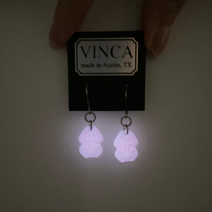 Pink penis earrings on stainless steel findings are pierced through a black branded Vinca card and are shown glowing in the dark. 