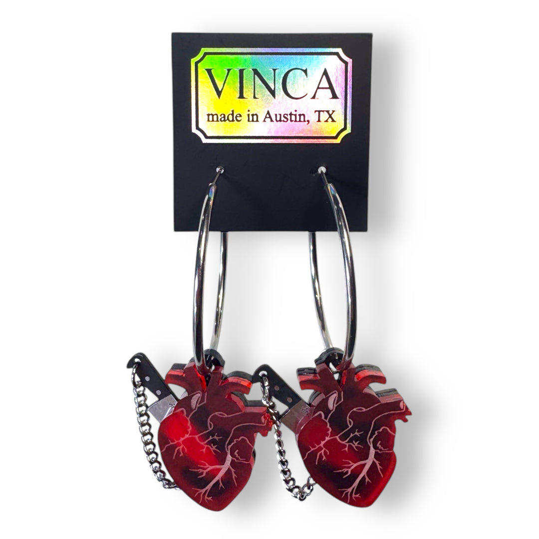 Red anatomical heart hoop earrings with removable chef's knife is on a black branded Vinca card set against a white background.