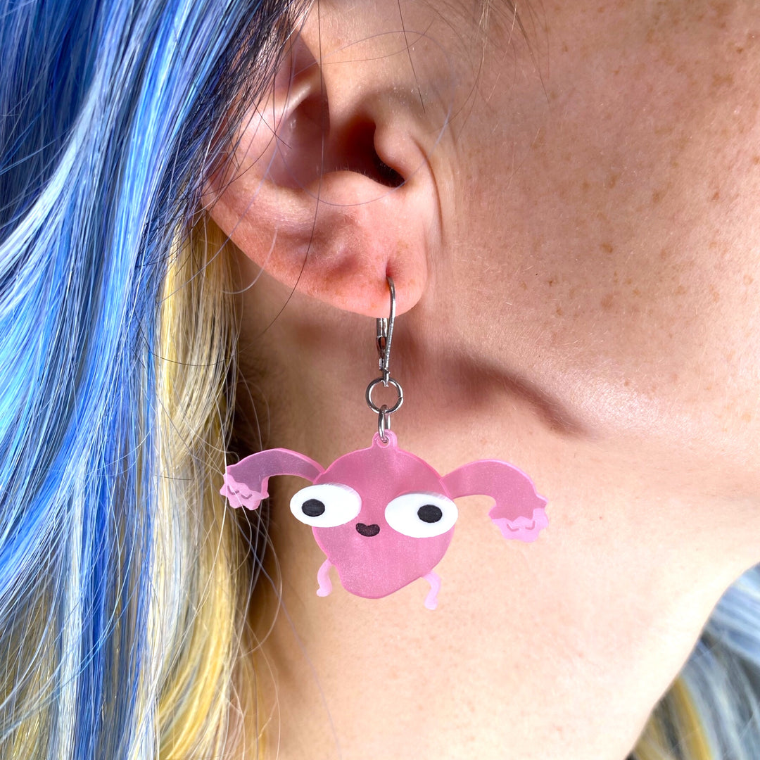 A woman with bright blue hair wears a Sarah's Scribbles Uterus dangle earring.