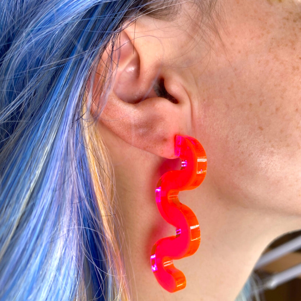 A woman with bright blue hair wears our Squiggle earrings in transparent neon pink.