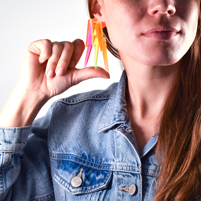 A woman showcases three different colors of lighting bolt earring colors including neon pink, neon green and neon orange. 