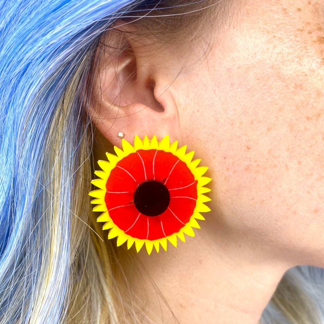 A large Indian Blanket wildflower earring is worn by a woman with bright blue hair.