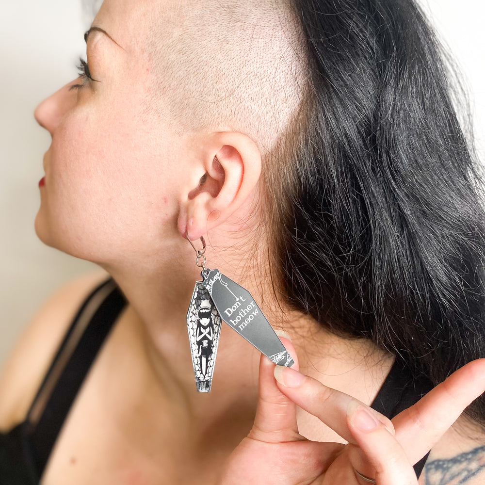 An alternative-goth model wears officially licensed Emily the Strange "Don't Bother Meow" stainless steel hook coffin earrings set against a white background.