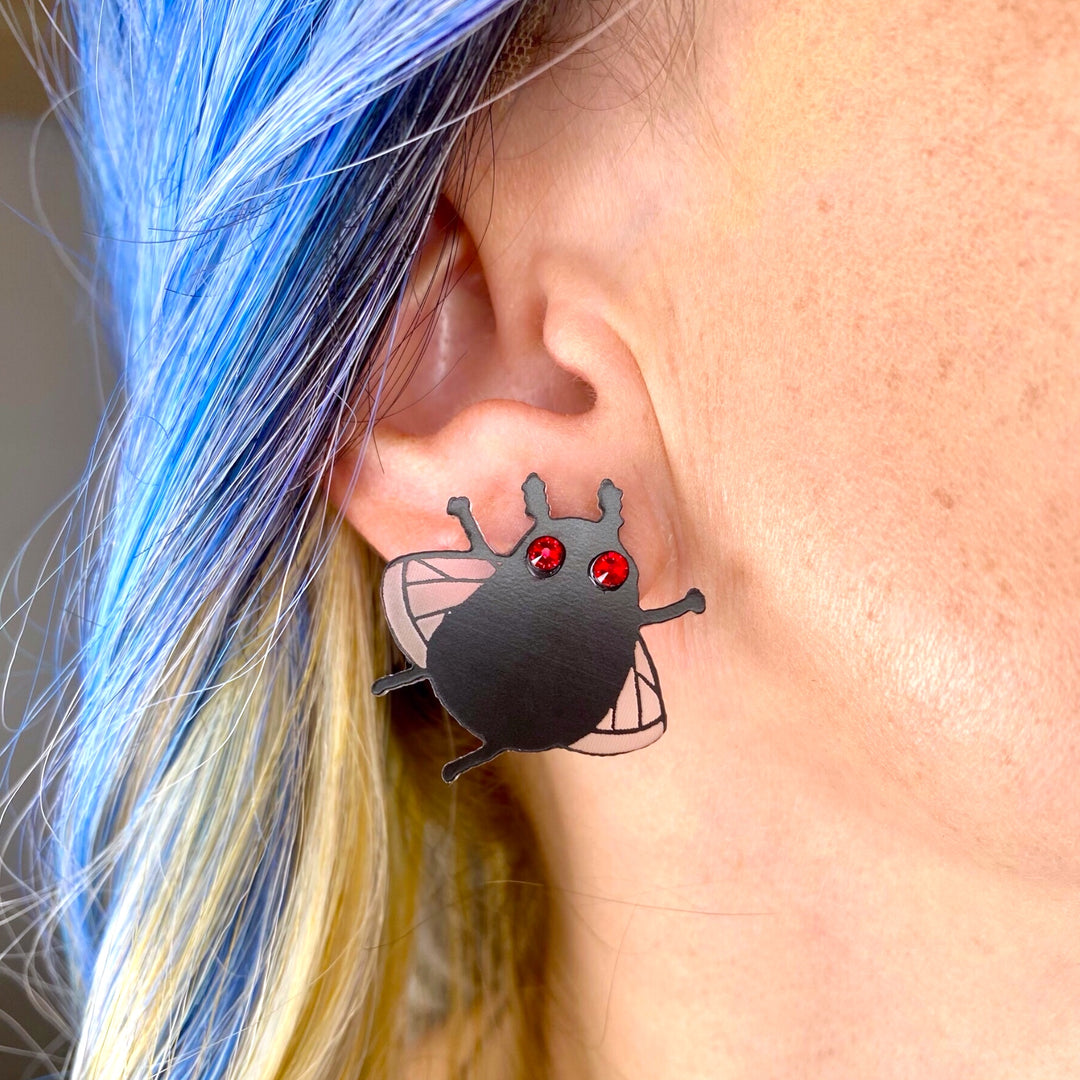 Our handmade Sarah's Scribbles Cryptid Club Moth Man stud earrings are worn by a model with blue hair.