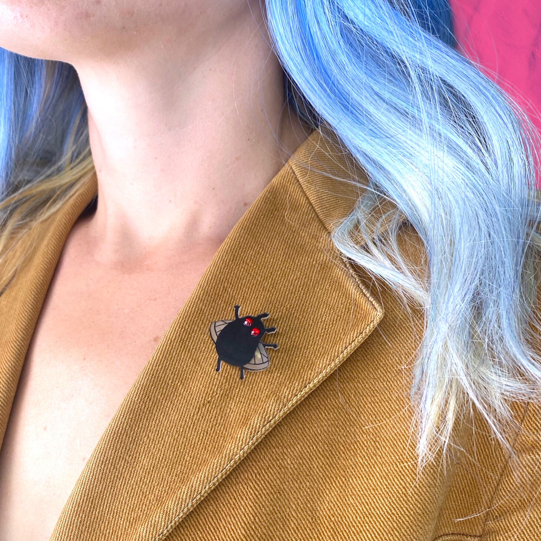 A model with blue hair wears our Sarah's Scribbles Cryptid Club Moth Man pin on the lapel of her tan blazer. 