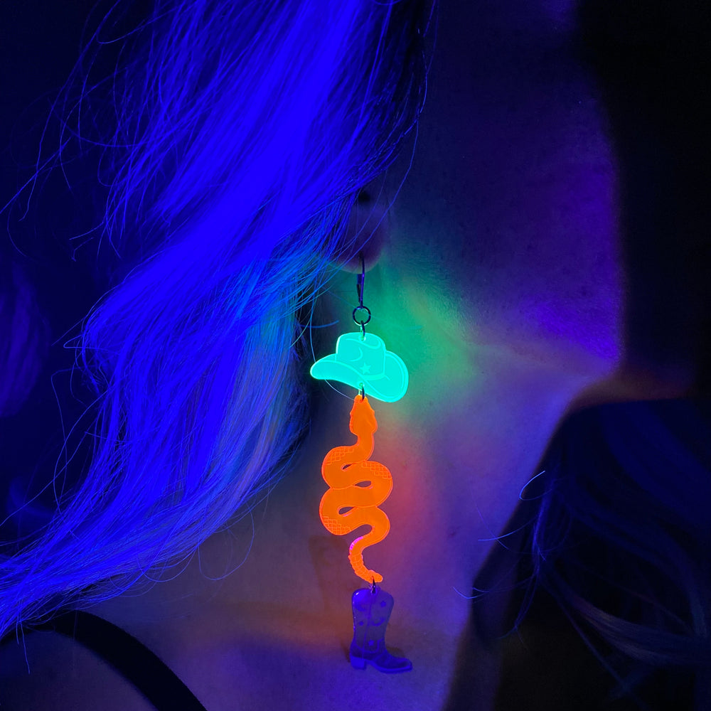 A woman wears a neon earring with a snake, cowboy hat and boot design glowing under black light.
