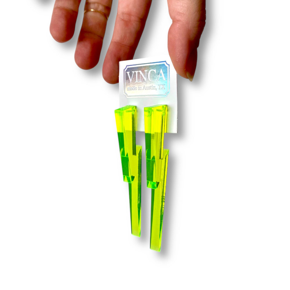 A hand holds a neon green lighting bolt earrings on a white Vinca branded card.