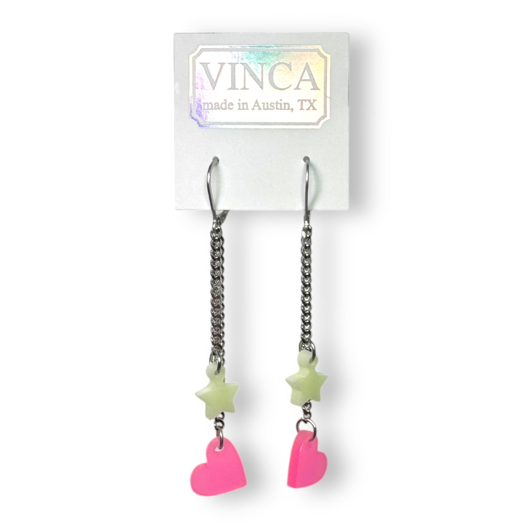 A pair of glow-in-the-dark pink heart and light green star stainless steel dangle earrings are attached to a white Vinca branded card.