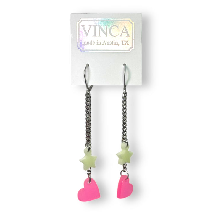 A pair of glow-in-the-dark pink heart and light green star stainless steel dangle earrings are attached to a white Vinca branded card.