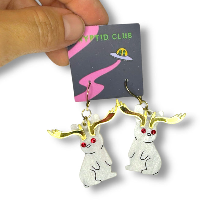 A hand holds a pair of Sarah's Scribbles Cryptid Club Jackalope Earrings on a branded card set against a white background.