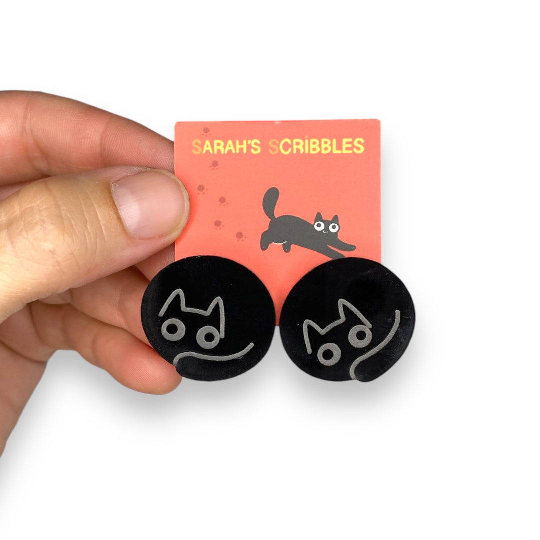 Our handmade Sarah's Scribbles Round Cat stud earrings in solid black are held over a white background.