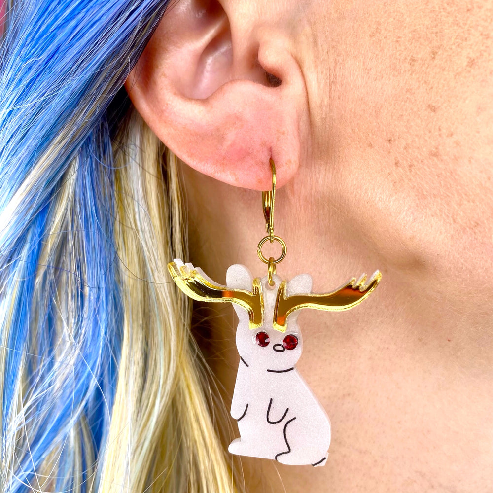 Sarah's Scribbles Cryptid Club Jackalope earrings are worn by a model with blue and platinum blonde hair.