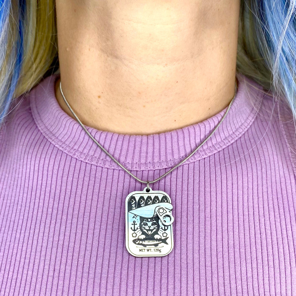 Person wearing a necklace with a tinned fish pendant against a ribbed lavender shirt.