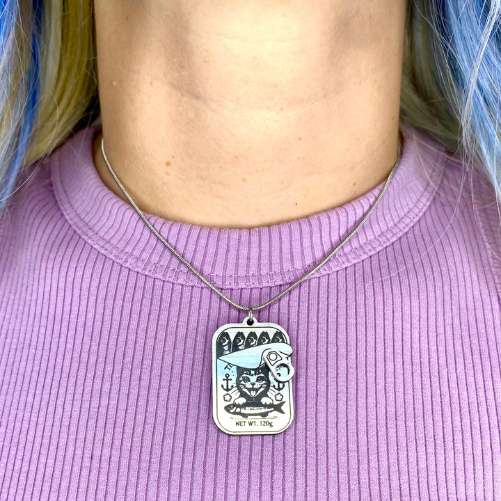 Person wearing a necklace with a tinned fish pendant against a ribbed lavender shirt.