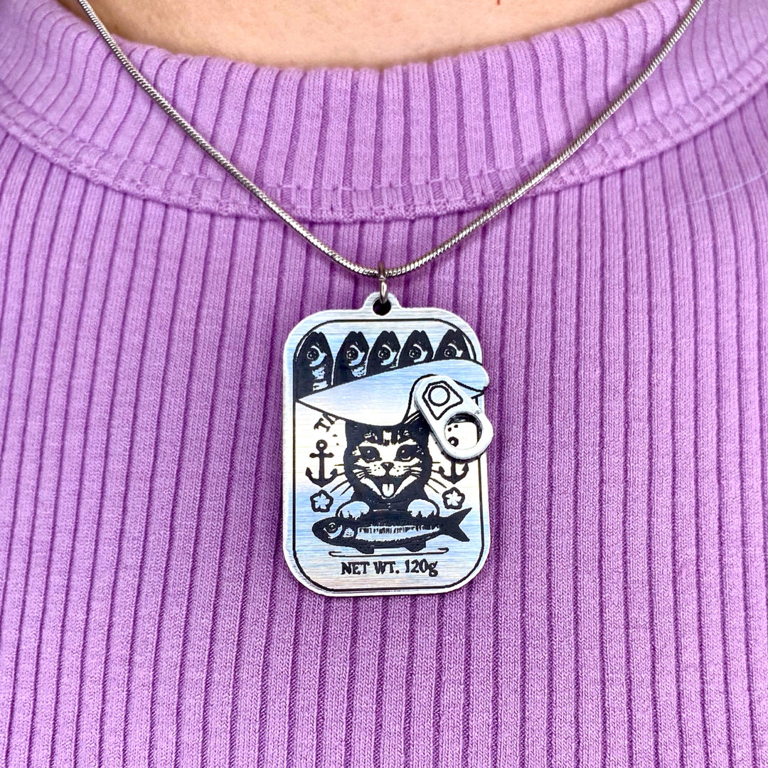 A silver-tone necklace with a charm depicting a kitten with a fish can, surrounded by anchors, worn on a purple ribbed garment.