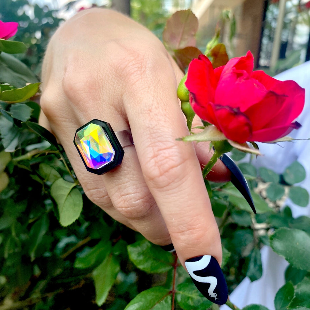 A hand wearing our adjustable Sorceress's Apprentice  Ring with a multicolored crystal centerpiece, set against a rose bush background.