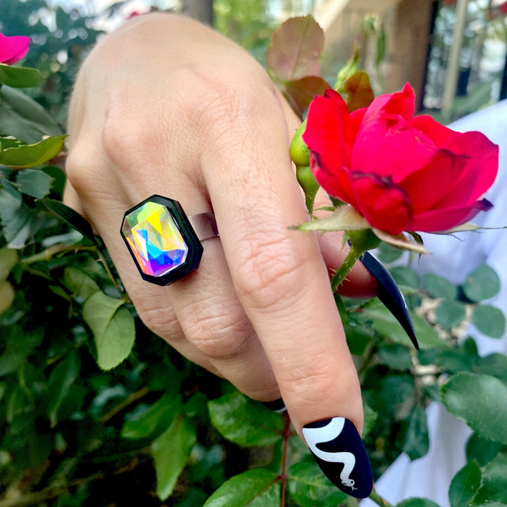 A hand wearing our adjustable Sorceress's Apprentice  Ring with a multicolored crystal centerpiece, set against a rose bush background.