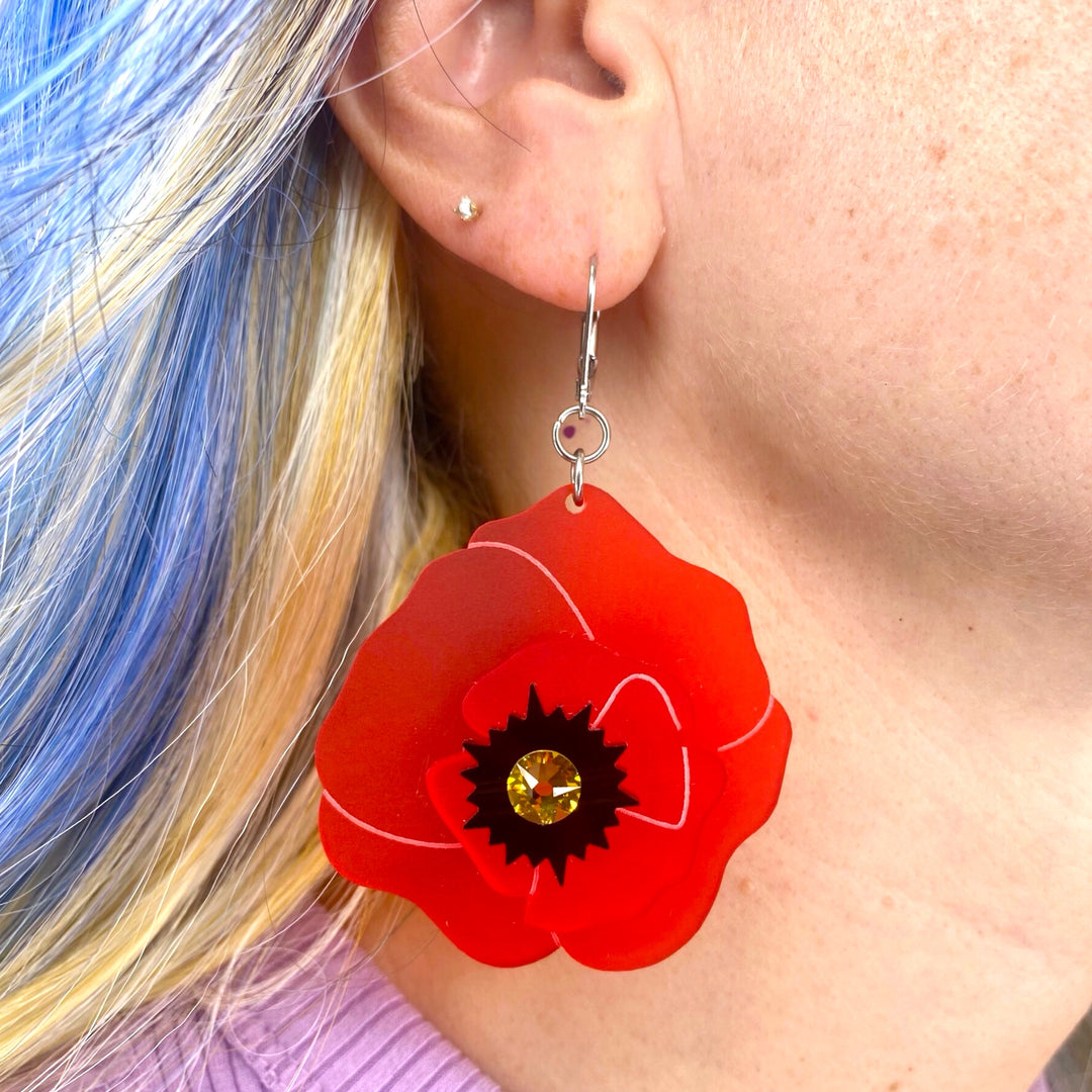 Bright red wildflower Poppy Dangle Earrings are worn by a woman with blue hair.
