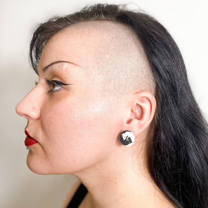 An edgy model wears our officially licensed Emily the Strange Yin Yang Cat glow-in-the-dark stud earrings by a white background.