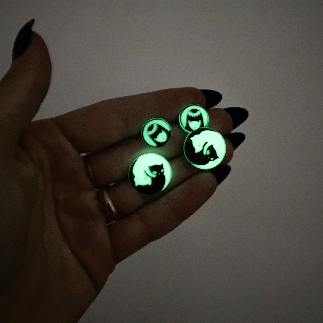 In the dark, a hand holds our officially licensed Emily the Strange Yin Yang Cat glow-in-the-dark stud earrings against a white background.