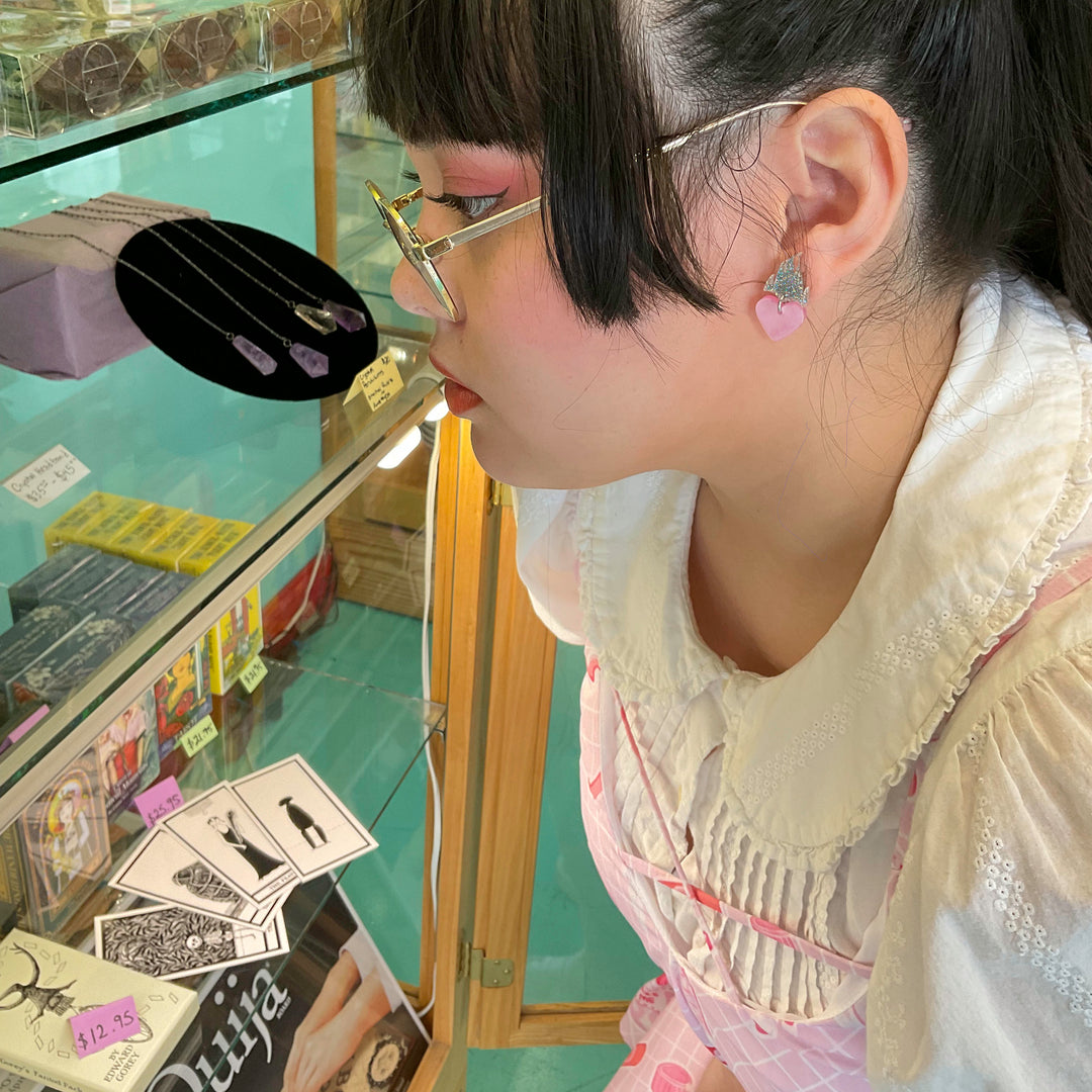 A young woman wears our flaming heart earrings in a pearl pink and glitter hologram in a toy store. 