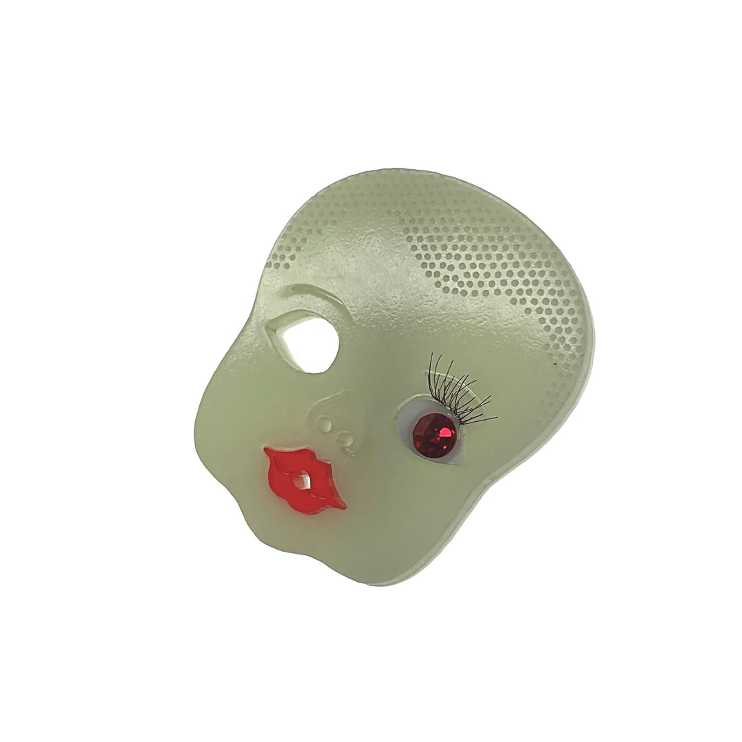 A brooch shaped like a creepy doll's head with a glow-in-the-dark head body and a crystal red eye, attached to a sturdy pinback.