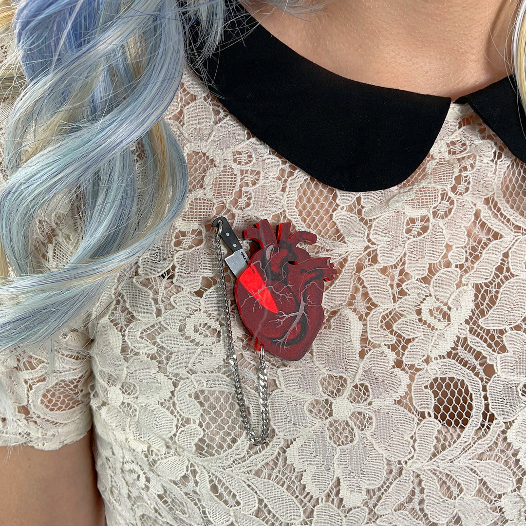 A woman with blue hair wearing a beige lace top with a black collar wears the heart of aghast brooch.