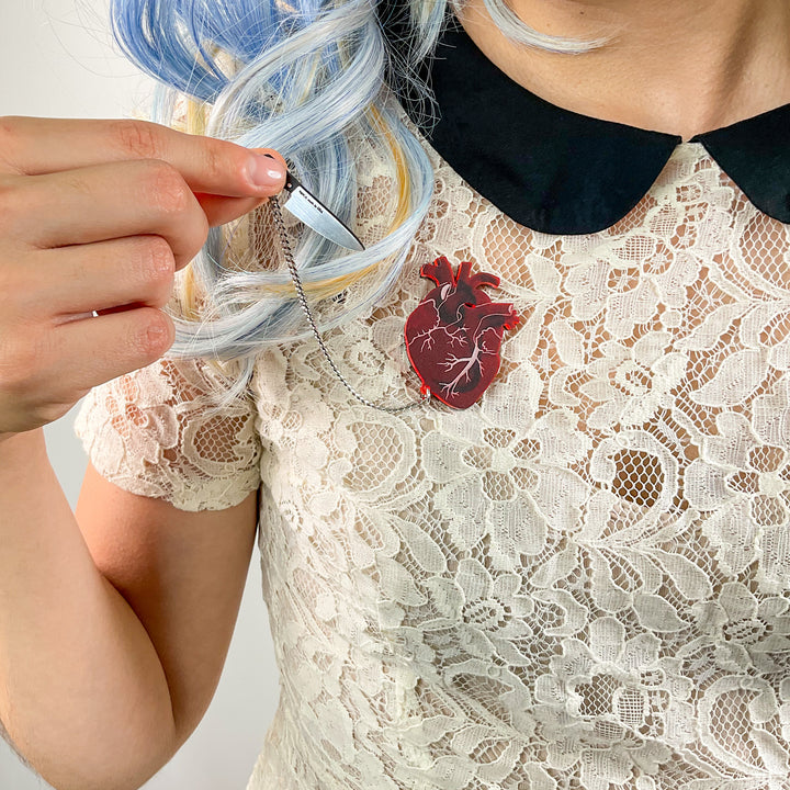 A woman wearing a cream colored lace top with a black collar wears an red anatomical heart brooch with a detachable knife.