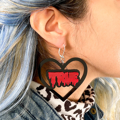 Person with colorful hair and a cheetah print top wears a black heart earring with red text that reads "true"