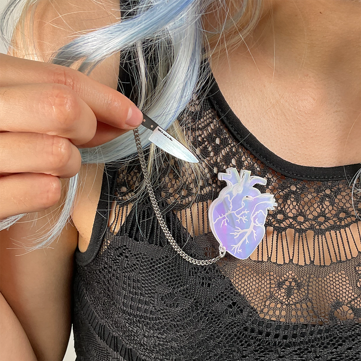 A woman with light blue hair wears Vinca's anatomical heart brooch with removable knife on her black lace top. She is holding the removable knife in her right hand.